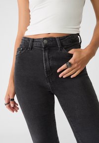 Mujer vestida con pantalones vaqueros de tiro alto negros y una camiseta corta blanca, con anillos de plata en ambas manos, uno con forma de rosa.