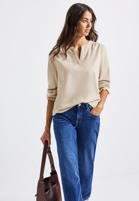 Frau in beiger Bluse mit langen Ärmeln und blauen Jeans, die eine dunkelbraune Lederhandtasche trägt und seitlich gegen einen weißen Hintergrund schaut.
