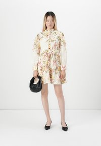 Donna che indossa un abito lungo a maniche lunghe con una cintura in vita, tiene una borsa nera e indossa delle scarpe nere con punta, il tutto su uno sfondo bianco.