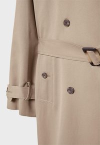 Trench coat beige con design doppiopetto, bottoni marroni, una cintura con passante e una tasca laterale con cuciture visibili.