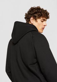 Svart hettegenser med teksturert stoff, med standard hette og en avslappet passform. Designet inkluderer synlige sømmer og en glatt finish.