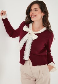 Burgundské svetlo s krajkovou mašlí, vroubkovaným krajkovým okrajem na límci a manžetách a ribbeným texturováním na celém materiálu.