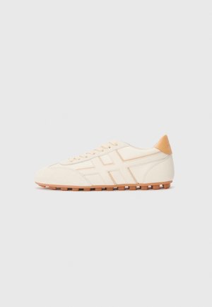 Low-profile beige sneaker with tan accents, lace-up front, textured rubber sole, and stitched cross pattern on the side.