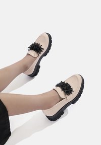 Scarpe slip-on beige con un accento decorativo a pom-pom nero, cuciture bianche a contrasto e suole in gomma nera spessa. Materiale liscio.