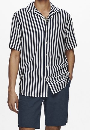 Uomo che indossa una camicia a maniche corte a righe verticali bianche e nere con pantaloncini blu navy, una mano in tasca.