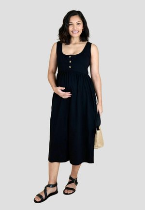 Nėščia moteris šypsosi, dėvi juodą midi suknelę be rankovių su sagomis, juodus sandalys ir laiko pintinę rankinę.