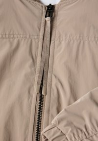 Veste beige légère avec une fermeture éclair centrale à dents noires et un tirette texturé ; manches légèrement froncées pour un effet modelant.