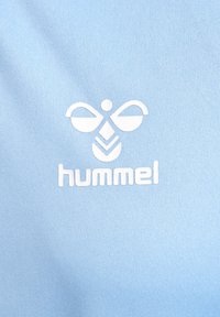 Logotipo blanco de Hummel con abeja estilizada y texto sobre fondo de tela texturizada azul claro.