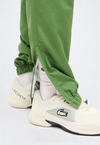 Λευκά αθλητικά παπούτσια Lacoste, φορεμένα με λευκές κάλτσες και πράσινο παντελόνι με πλαϊνά φερμουάρ και ελαστικές μανσέτες.
