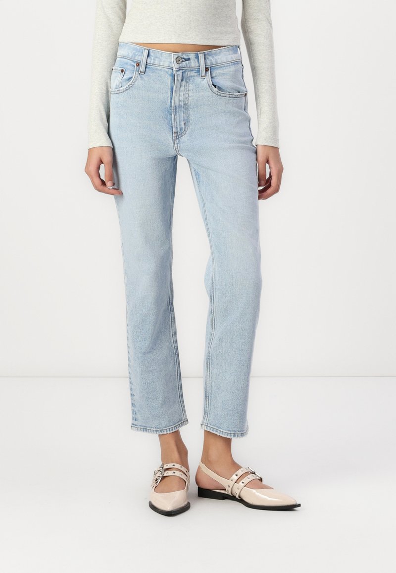 Jeans droits bleu clair avec taille haute, dotés de deux poches avant et d'une fermeture à boutons, accompagnés de chaussures à bride arrière beiges.