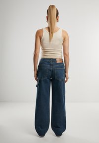 Plačios, tamsiai mėlynos spalvos džinsinės kelnės su aukštu liemeniu, daug kišenių ir minkšta tekstūra, derinamos su aptemptu smėlio spalvos marškinėliais be rankovių.