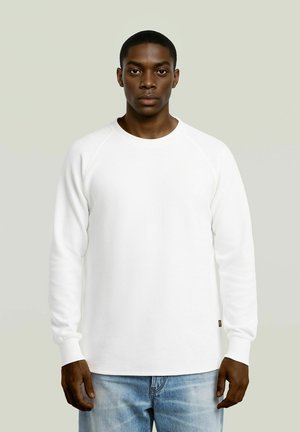 Junger Mann mit dunkler Hautfarbe, der ein schlichtes weißes Langarmshirt und hellblaue Jeans trägt, steht vor einem einfarbigen Hintergrund und schaut nach vorne.