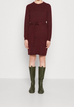 Osoba ubrana w bordową sweterkową sukienkę długości do kolan z paskiem w talii oraz oliwkowo-zielone, masywne kozaki do kolan na jednolitym tle.