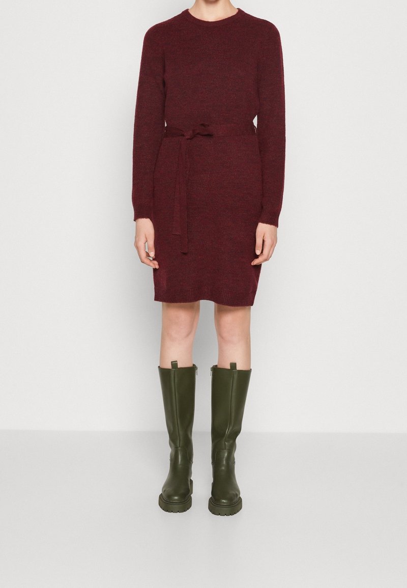 Personne portant une robe tricotée bordeaux mi-genou avec une ceinture à la taille et des bottes épaisses kaki montant jusqu'au genou, sur un fond uni.