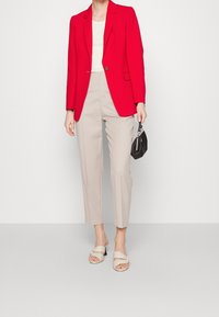 Donna con blazer rosso, top bianco, pantaloni beige a cropped, sandali beige con tacco, che tiene una piccola borsa nera su sfondo bianco.