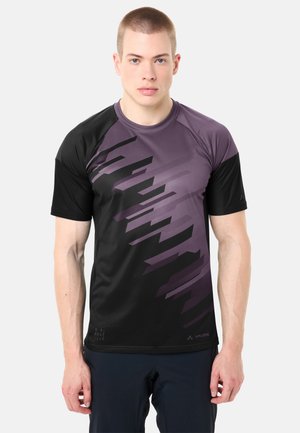 Giovane uomo con capelli corti che indossa una maglia sportiva nera con motivo geometrico viola e pantaloni scuri, in piedi contro uno sfondo semplice.