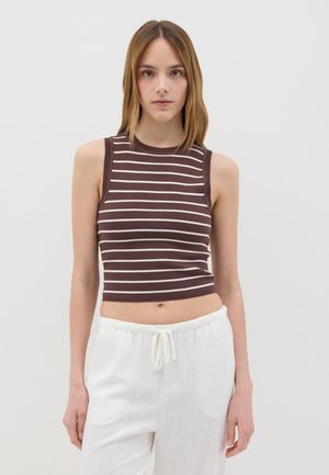 Top corto senza maniche a righe marrone e crema con una texture a costine, caratterizzato da un collo alto. Abbinato a pantaloni bianchi leggeri e morbidi.