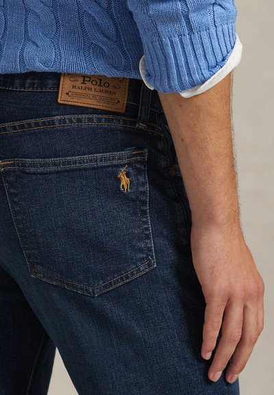 Približna snimka osobe koja nosi tamnoplave traperice s logotipom Polo Ralph Lauren i plavi džemper s kabelastim uzorkom s bijelim manžetnama košulje.