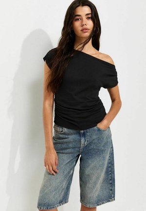 Jonge vrouw met lang haar, gekleed in een zwart off-shoulder top en losse denim shorts, staat met één hand in de zak.