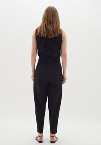 Top senza maniche nero con schiena arricciata, abbinato a pantaloni neri a vita alta che si stringono alla caviglia; modella che indossa sandali neri.