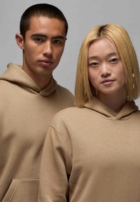 Due persone indossano felpe con cappuccio beige, dotate di tasca frontale e polsini a coste, su uno sfondo grigio uniforme.