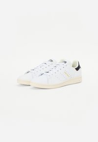 Hvide lædersneakers med perforerede detaljer, flade snørebånd, et gult logo og en kontrasterende sort og orange hælkappe. Gummisål.