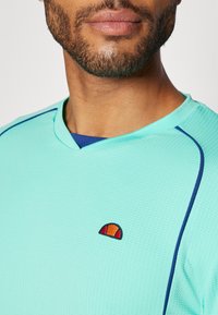 Light teal short-sleeve athletic shirt with textured fabric, v-neckline, blue accents, and a small orange and red logo on the chest.