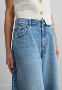 Femme portant un jean en denim bleu clair taille haute et un haut blanc sans manches avec des boutons dorés, debout devant un fond uni.