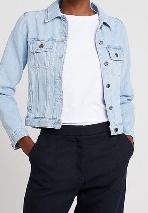 Personne portant une veste en jean bleu clair par-dessus un t-shirt blanc, les mains dans les poches d'un pantalon noir taille haute.