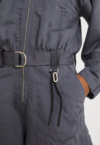 Gros plan d'une combinaison gris foncé à fermeture éclair, avec une ceinture en tissu et une boucle métallique, un détail de cordon noir et une main dans la poche latérale.