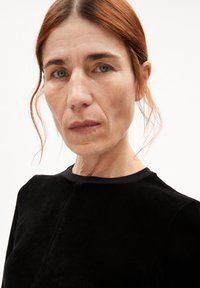 Zwarte fluwelen top met een ronde halslijn, voorzien van een subtiele naaddetail aan de voorkant en een zachte textuur. Haar in losse golven gestyled.