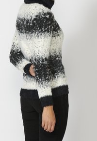 Person in einem schwarz-weißen, gemusterten Rollkragenpullover und schwarzen Hosen, mit der Hand auf dem unteren Rücken, vor einem schlichten Hintergrund.