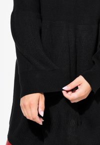 Maglione nero lavorato a maglia con texture a costine, maniche ampie e vestibilità morbida. Le mani sistemano l'orlo, mettendo in evidenza il design rilassato del capo.