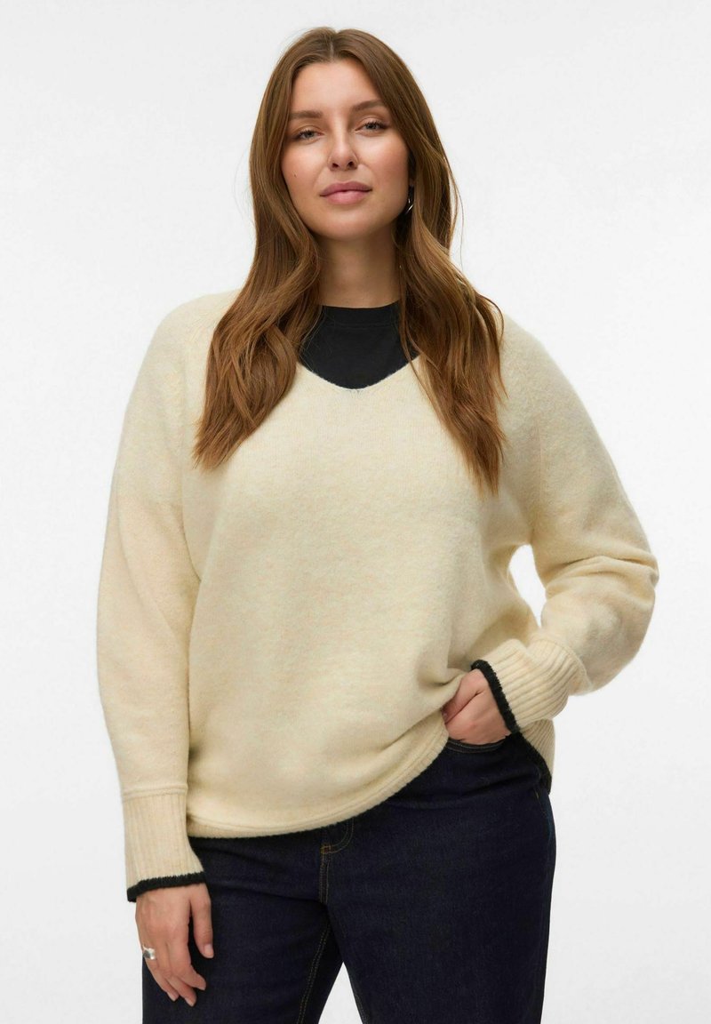 Femme avec de longs cheveux bruns portant un pull crème sur un haut noir et un jean foncé, posant avec une main dans la poche sur un fond blanc.