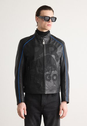 Black leather jacket with embossed branding, blue accent stitching, high collar, and zip closure. Worn over a black turtleneck.