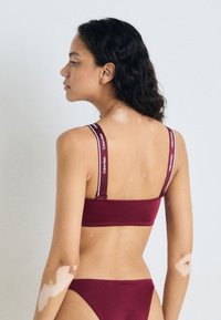 Maroon bralette s podesivim trakama s bijelim brendingom, u kombinaciji s odgovarajućim badić donjim dijelom. Glatka tekstura i prilagođeni dizajn.