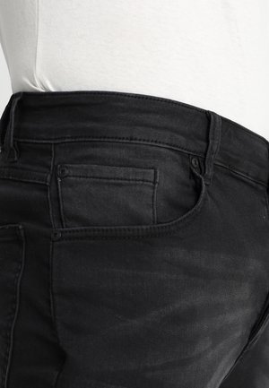 Close-up van een persoon die zwarte spijkerbroek draagt met een voorzak en riemlussen, gecombineerd met een wit, ingestopt shirt.