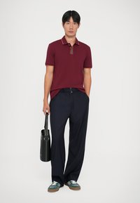 Κερασί μπλουζάκι πόλο με ριγωτό γιακά, συνδυασμένο με ναυτικά ριγωτά παντελόνια φάρδους. Μαύρη τσάντα στο χέρι και πράσινα αθλητικά παπούτσια.