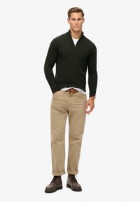 Maglione verde scuro con zip al collo, realizzato in tessuto a maglia, abbinato a pantaloni chinos beige e stivali marroni con lacci. Presenta una vestibilità casual e rilassata.