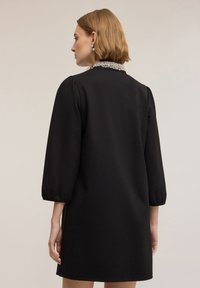 Czarna sukienka o dopasowanym kroju, z ozdobnym kołnierzem wysadzanym koralikami oraz rękawami o długości trzy czwarte z bufkami. Gładka faktura, długość do kolan.