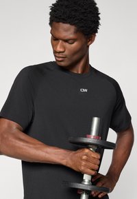 Μαύρο αθλητικό κοντομάνικο μπλουζάκι με στρογγυλή λαιμόκοψη, φορεμένο από άτομο που κρατάει αλτήρα. Η μπλούζα φέρει λογότυπο στο πάνω αριστερό μέρος.