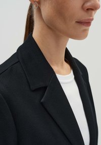 Zwarte gemarmerde blazer met een gesneden kraag, gedragen over een witte top, met een gladde stof en een close-upweergave van de kraag en oordetail.