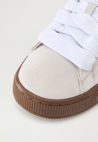 Sneaker med en beige semsket overdel, hvite vevde lisser og en teksturert brun gummisåle. Rund tå med subtile sømdetaljer.