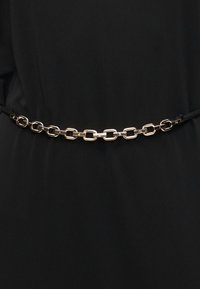 Ζώνη αλυσίδα σε χρυσό τόνο με ορθογώνιους κρίκους, φορεμένη πάνω σε μαύρο υφασμάτινο φόντο, στερεωμένη με λεπτό μαύρο λουρί σε κάθε πλευρά.