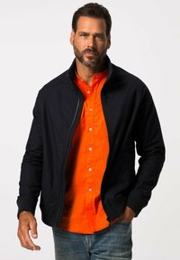 Svart zip-jacka med ribbade ärmar över en orange knapp-up skjorta. Blå jeans fulländar outfiten. Jackan har en slät textur.