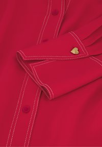 Heldere rode shirt met een puntkraag, zichtbare knopen en contrasterende witte stiksels. Bevat een klein gouden hartaccent bij de manchet.