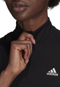 Černý sportovní top s vysokým límcem, zipovým zapínáním a malým bílým logem adidas na hrudi. Hladká tkanina s přiléhavým střihem.