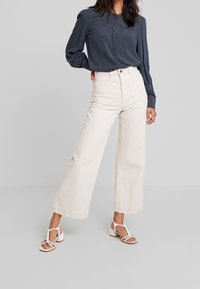 Vaaleansävyiset leveälahkeiset farkut korkealla vyötäröllä, yhdistetty tummansiniseen pitkähihainen pusero, jossa on pieni valkoinen pilkullinen kuvio. Valkoiset remmiballerinat.