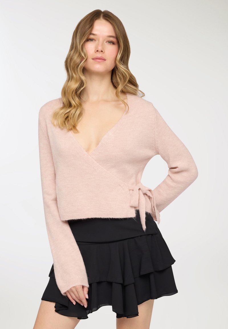 Maglione rosa, corto e avvolgente, realizzato con materiale morbido. Presenta una scollatura a V profonda e un dettaglio con fiocco laterale, abbinato a una gonna nera a strati.