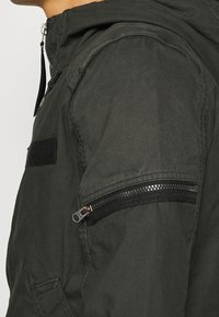 Mörkgrön huva jacka med en strukturerad tyg, dragkedja ficka på ärmen och justerbara dragsnören vid kragen.